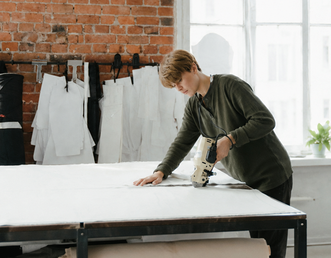 A person uses an electric rotary cutter to cut white fabric on a large table in a bright studio.